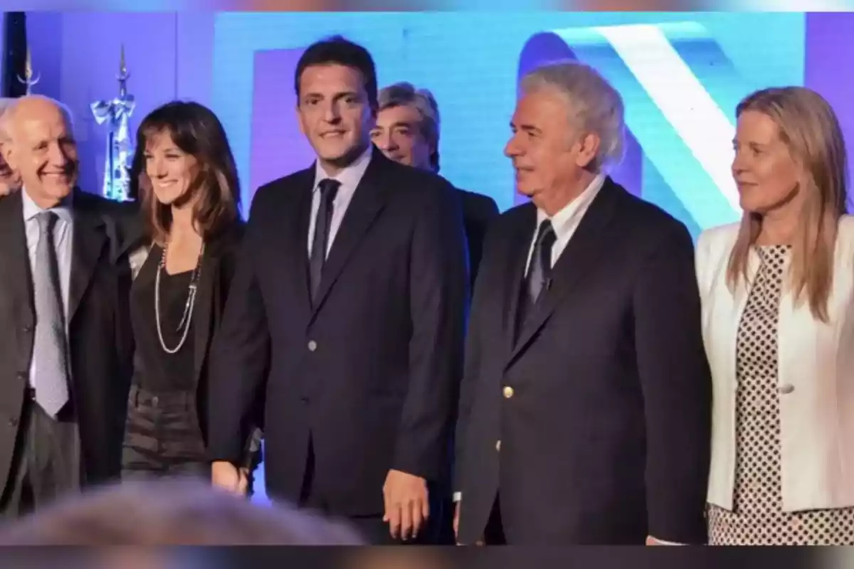 Cinco personas vestidas de manera formal posan juntas y sonríen en un evento con fondo azul Cinco personas vestidas de manera formal posan juntas y sonríen en un evento con fondo azul