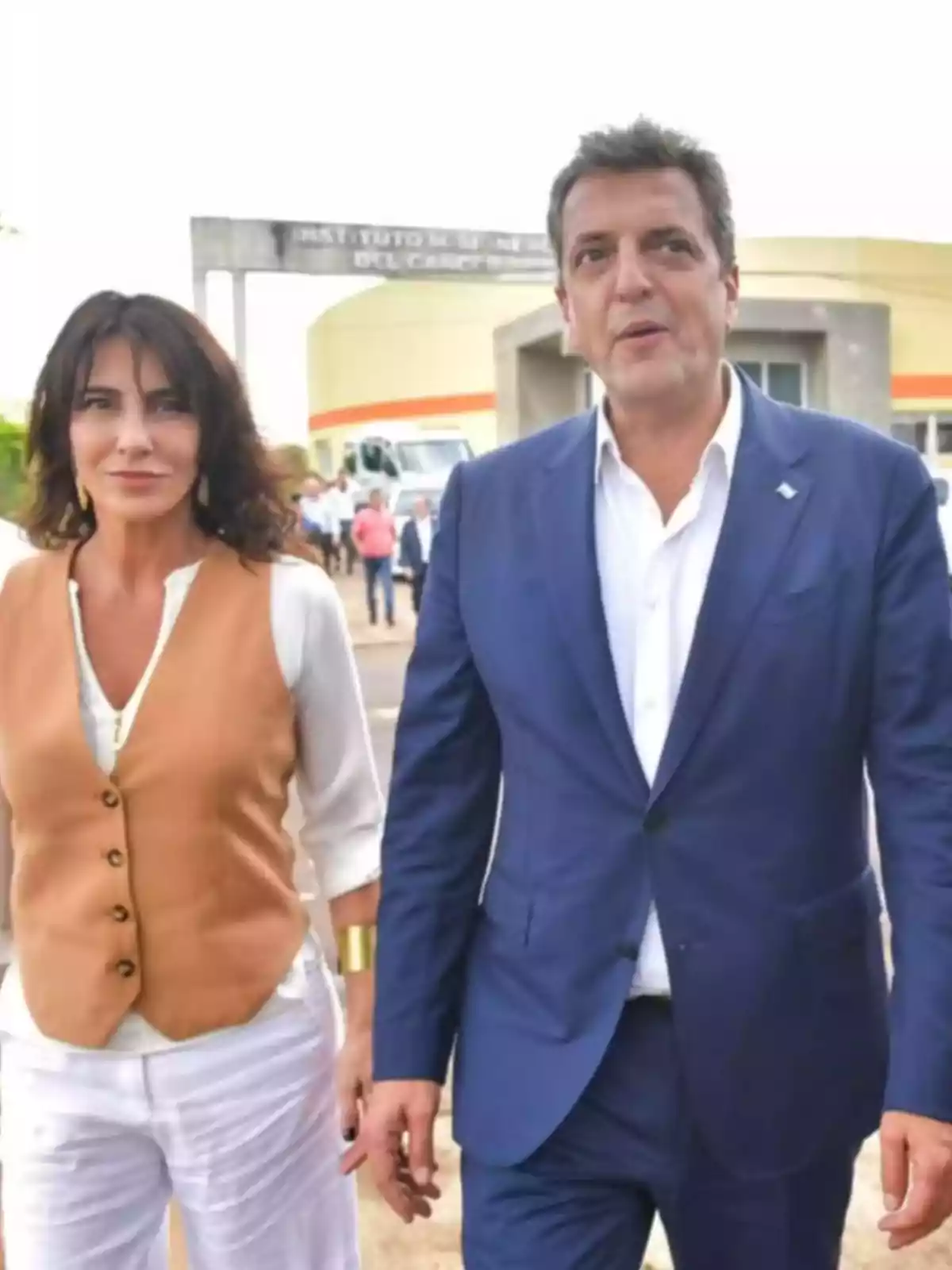 Una mujer con chaleco marrón y un hombre con traje azul caminan juntos al aire libre frente a un edificio con personas al fondo Una mujer con chaleco marrón y un hombre con traje azul caminan juntos al aire libre frente a un edificio con personas al fondo