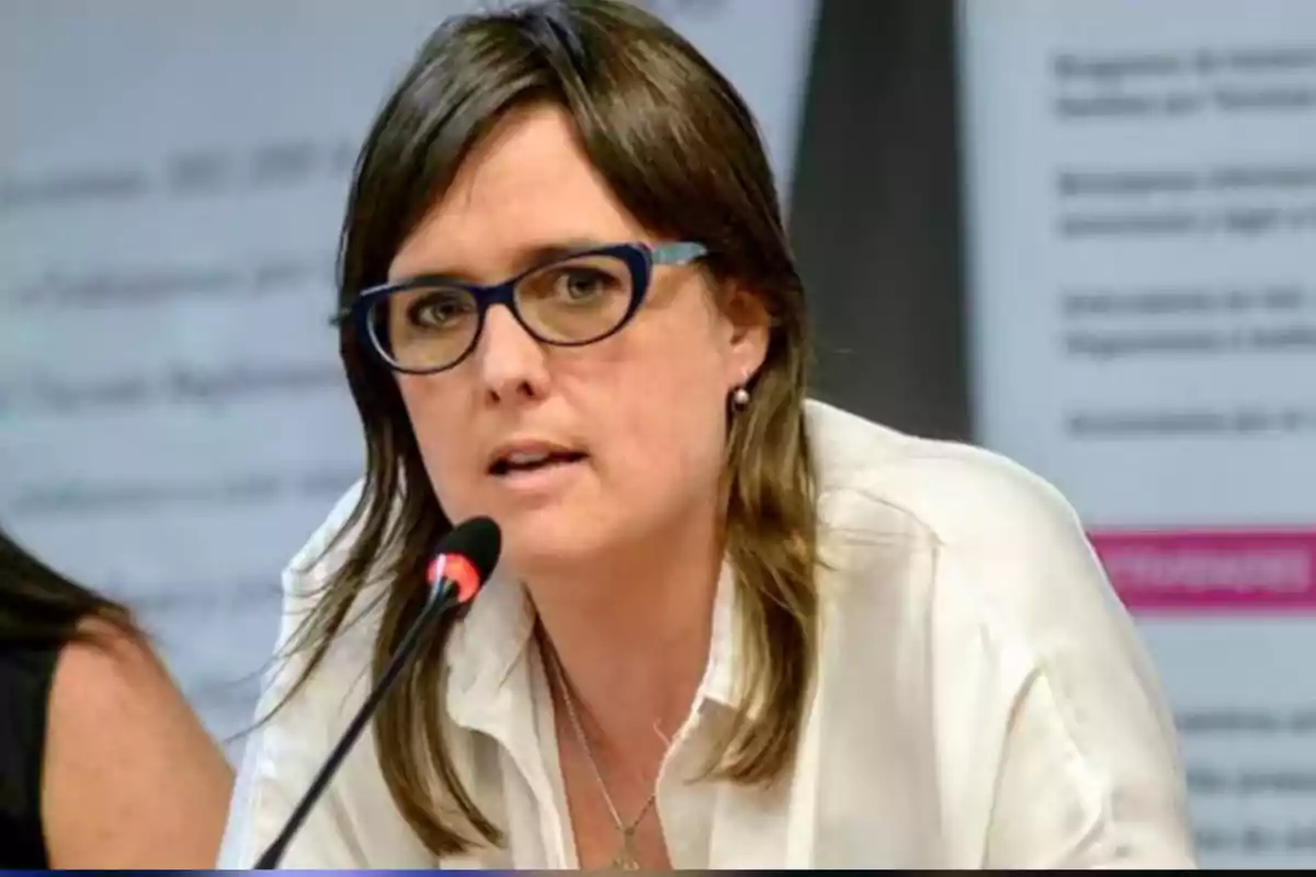 Mujer con gafas y camisa blanca hablando frente a un micrófono en un entorno profesional Mujer con gafas y camisa blanca hablando frente a un micrófono en un entorno profesional