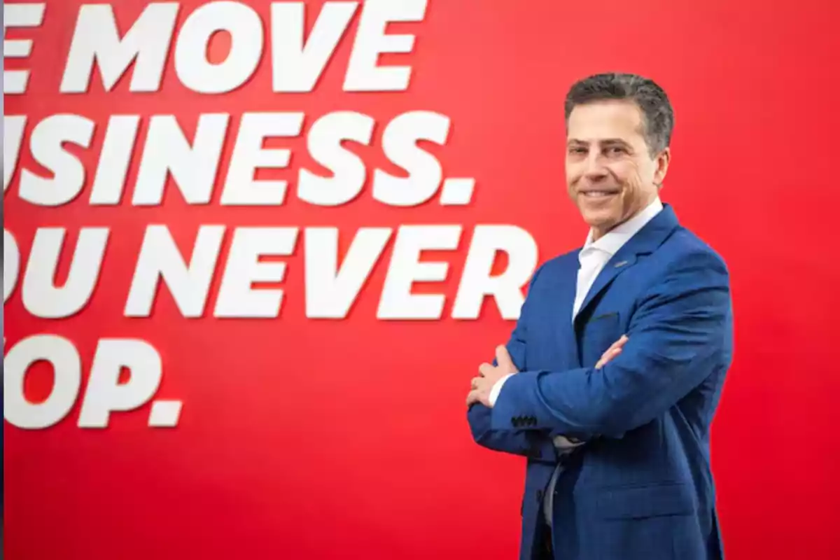Hombre de traje azul sonriendo con los brazos cruzados frente a una pared roja con texto en letras blancas Hombre de traje azul sonriendo con los brazos cruzados frente a una pared roja con texto en letras blancas