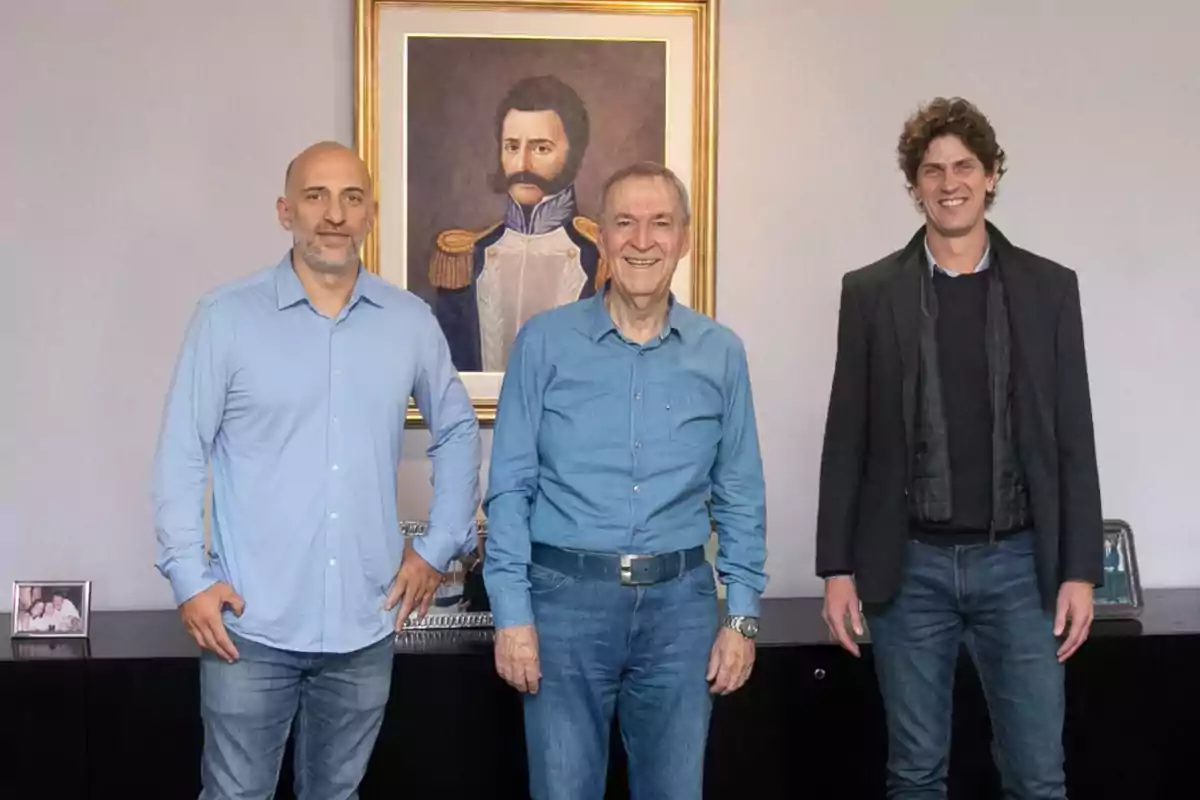Three men pose smiling in front of a painting of a soldier in an old-fashioned uniform in an office. Three men pose smiling in front of a painting of a soldier in an old-fashioned uniform in an office.