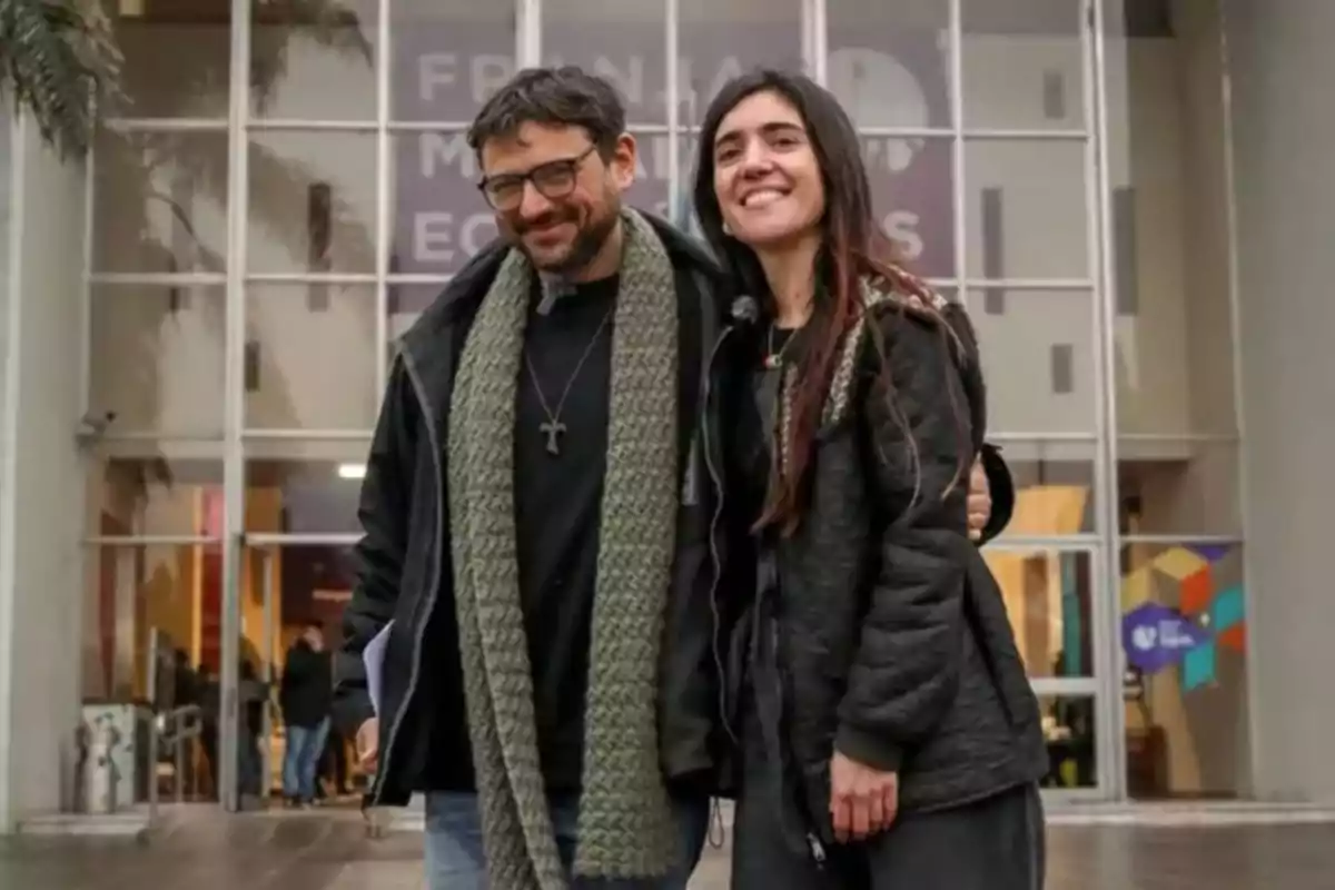Dos personas sonríen y posan abrazadas frente a un edificio de cristal, ambas llevan ropa de abrigo y se ven contentas. Dos personas sonríen y posan abrazadas frente a un edificio de cristal, ambas llevan ropa de abrigo y se ven contentas.