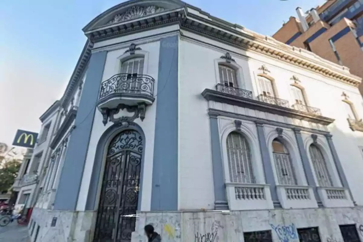 Old building with a white and blue facade, wrought iron balconies, and arched windows; in the background, a fast food restaurant sign can be seen.