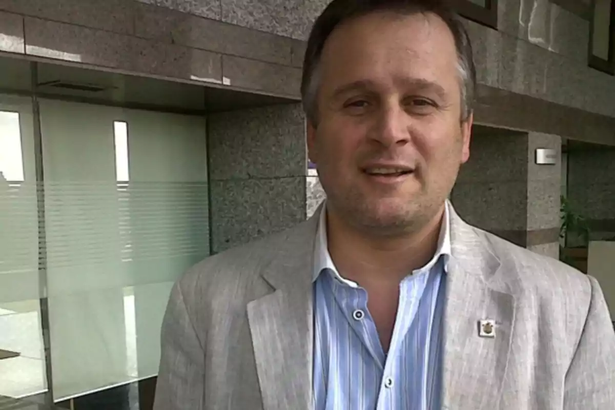 Hombre de cabello corto y canoso viste saco claro y camisa azul a rayas en un entorno de oficina con paredes de mármol y vidrio.