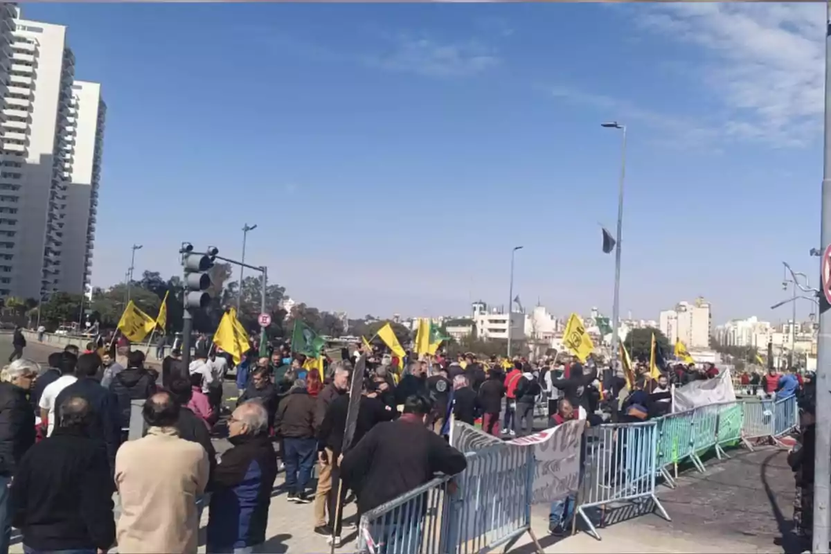 Un grupo de personas se manifiesta en la calle detrás de vallas de seguridad, algunas sostienen banderas amarillas y verdes, mientras al fondo se ven edificios altos y el cielo despejado. Un grupo de personas se manifiesta en la calle detrás de vallas de seguridad, algunas sostienen banderas amarillas y verdes, mientras al fondo se ven edificios altos y el cielo despejado.