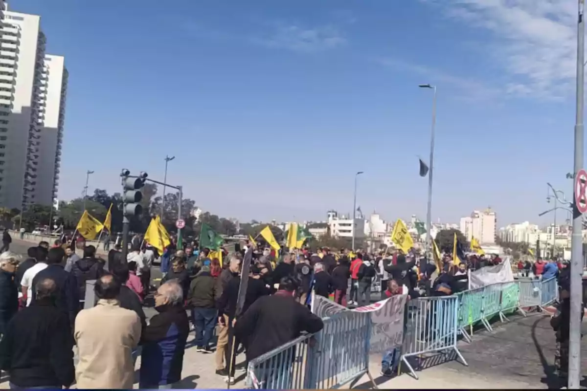 A group of people is protesting in the street behind security barriers, some holding yellow and green flags, while tall buildings and a clear sky can be seen in the background.