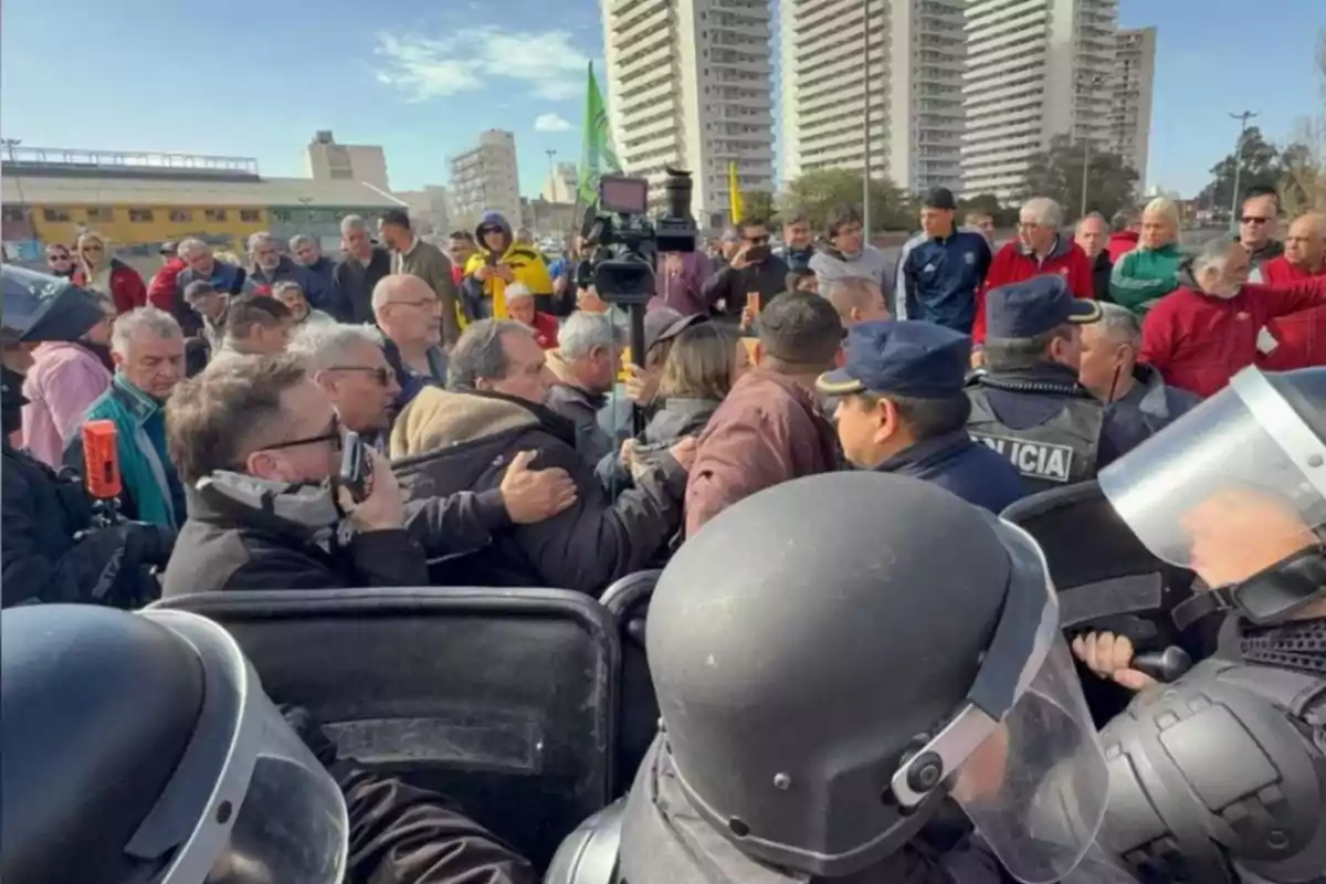 A group of people confronts riot police at an outdoor protest, with tall buildings in the background and several people watching the situation. A group of people confronts riot police at an outdoor protest, with tall buildings in the background and several people watching the situation.