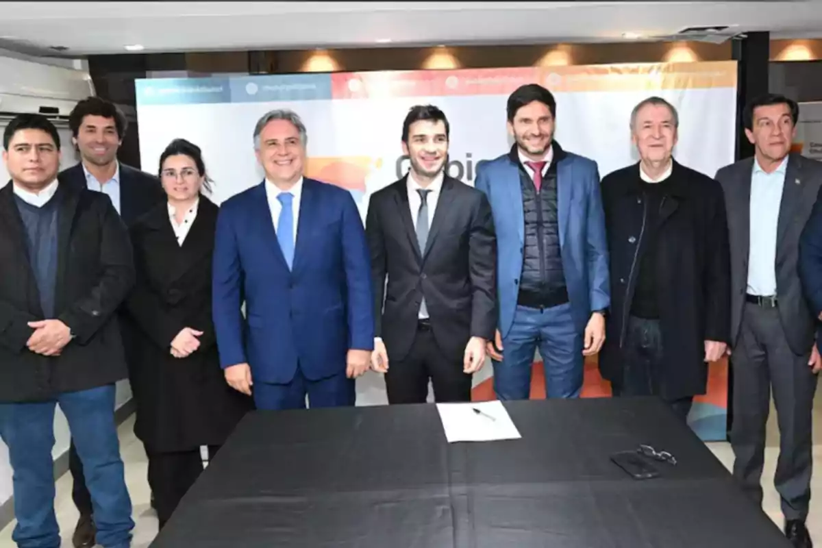 Grupo de personas vestidas de manera formal posando para una foto frente a una mesa con un mantel negro y un documento sobre ella, con un fondo de banner institucional. Grupo de personas vestidas de manera formal posando para una foto frente a una mesa con un mantel negro y un documento sobre ella, con un fondo de banner institucional.