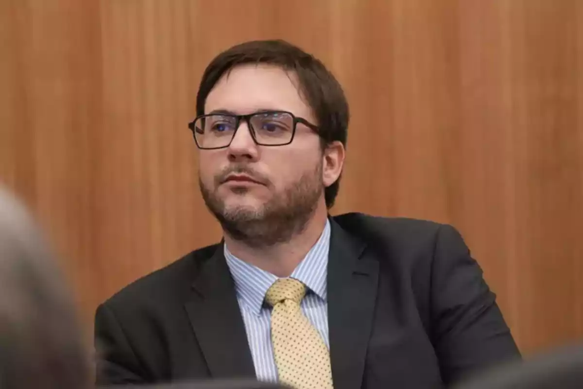 Man with glasses and a short beard wearing a dark suit, blue jersey, and yellow polka-dot tie, seated in front of a wooden background Man with glasses and a short beard wearing a dark suit, blue jersey, and yellow polka-dot tie, seated in front of a wooden background
