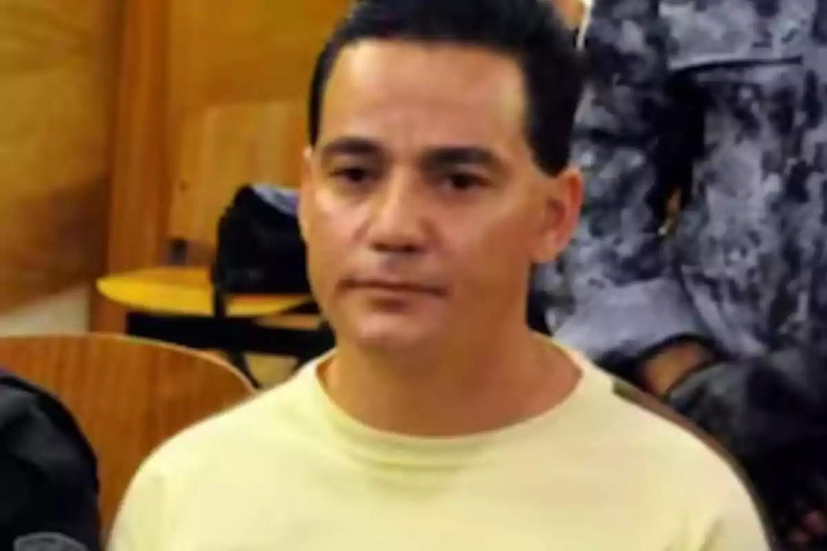 Short dark-haired man wearing a light-colored jersey sitting in a room with other people Short dark-haired man wearing a light-colored jersey sitting in a room with other people