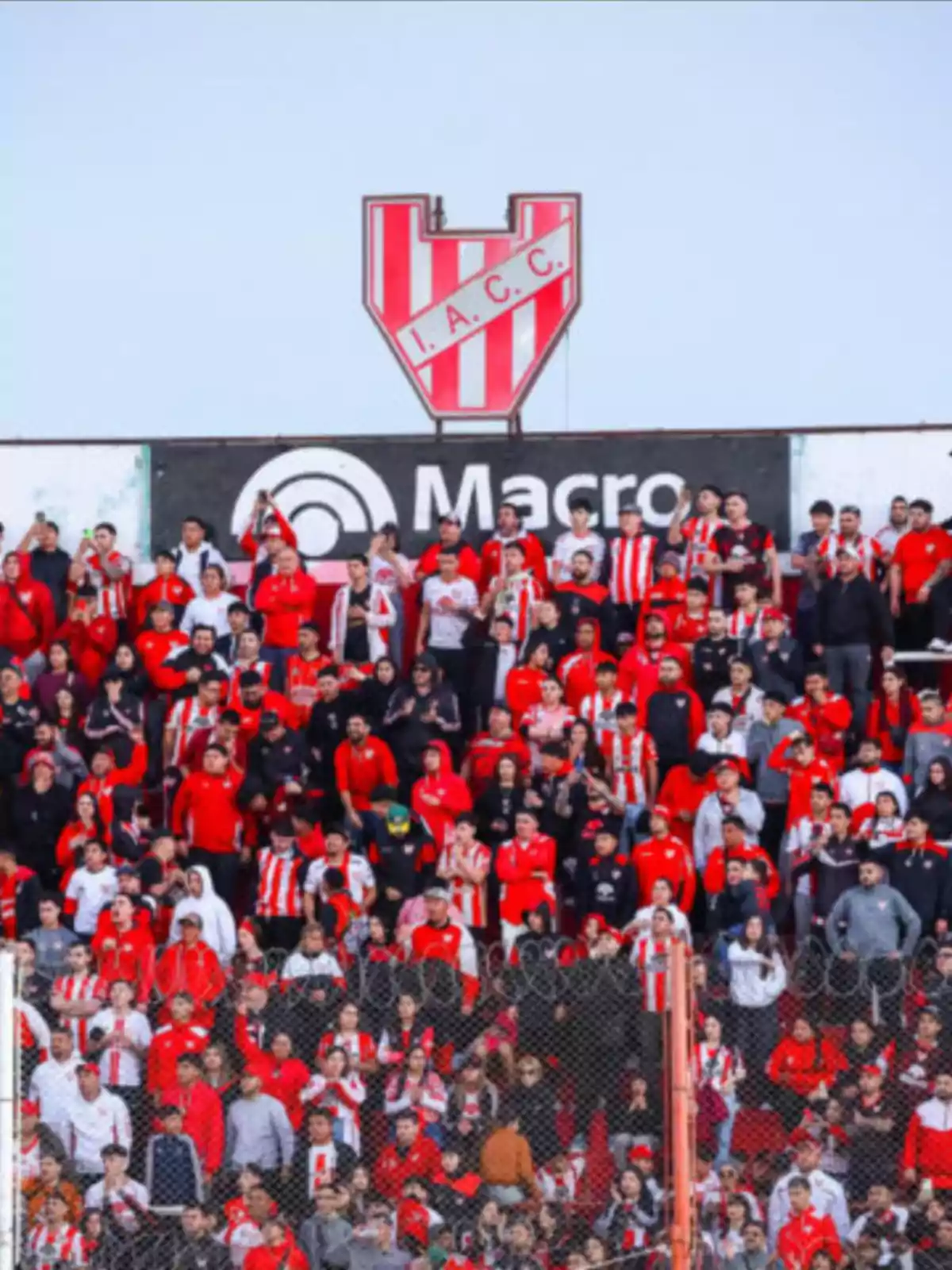 Fans dressed in red and white cheer in a stadium stand with a large Instituto Atlético Central Córdoba crest in the background. Fans dressed in red and white cheer in a stadium stand with a large Instituto Atlético Central Córdoba crest in the background.