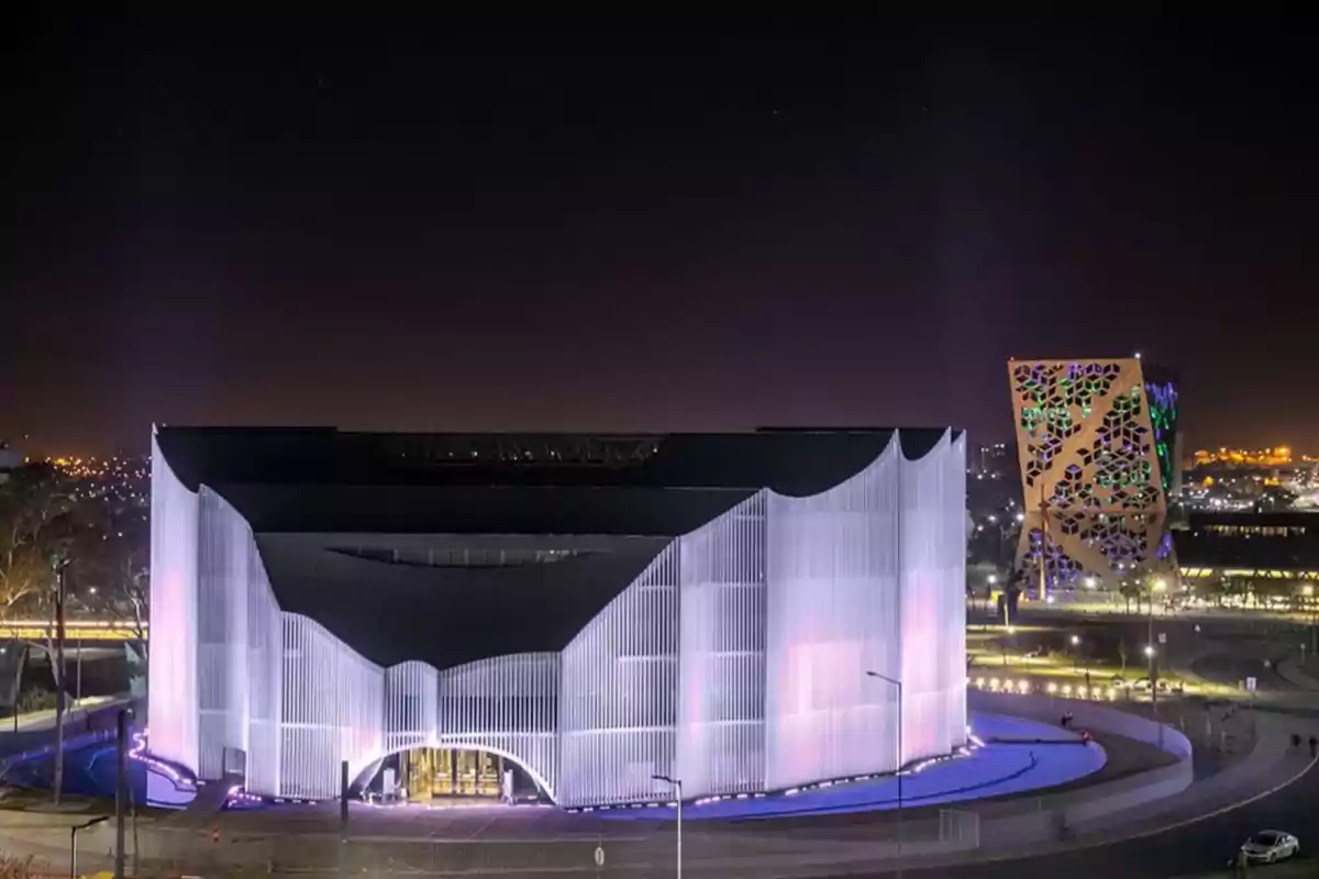 Modern building illuminated at night with a wavy facade and decorative structure in the background in a city