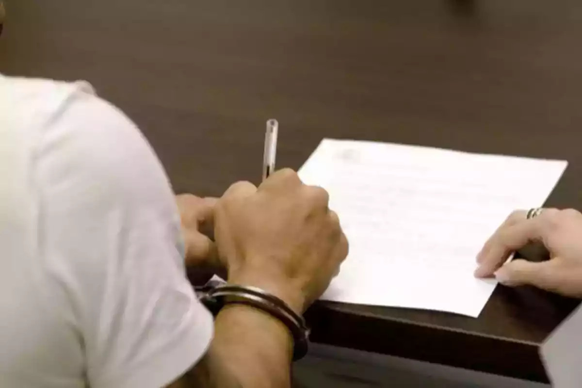 Persona esposada firmando un documento en una mesa junto a otra persona Persona esposada firmando un documento en una mesa junto a otra persona