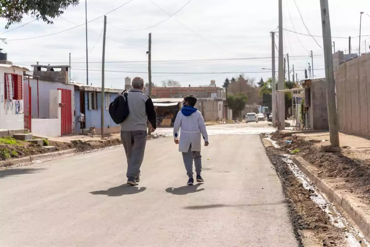 Un hombre y un niño caminan por una calle de un barrio humilde en un día soleado