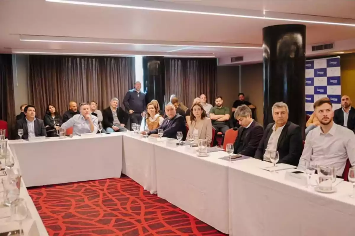 A group of people sitting around white tables in a meeting room with red carpet and dark curtains, paying attention to a presentation or talk. A group of people sitting around white tables in a meeting room with red carpet and dark curtains, paying attention to a presentation or talk.