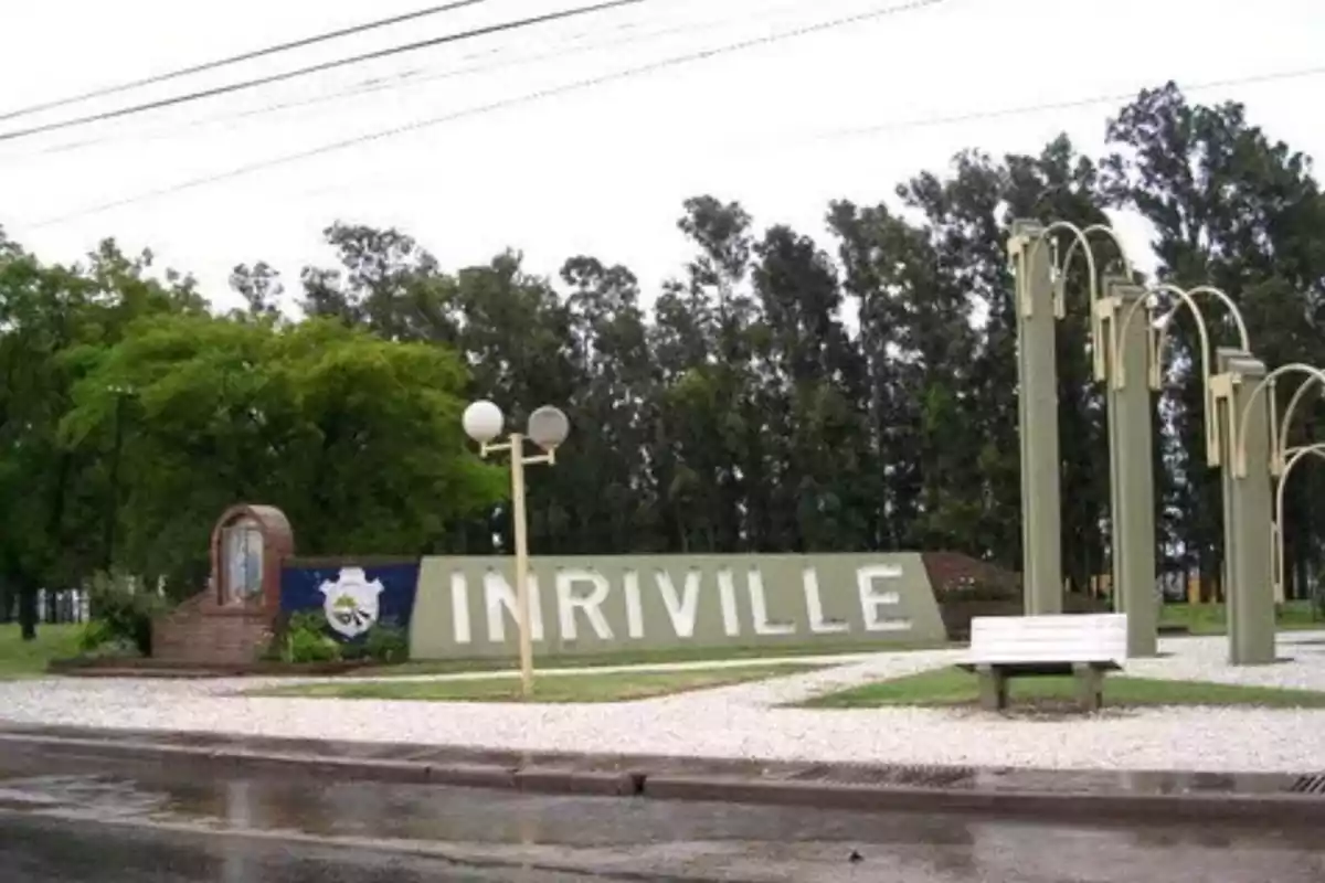 Welcome sign to Inriville surrounded by trees, streetlights, and a white bench in a plaza
