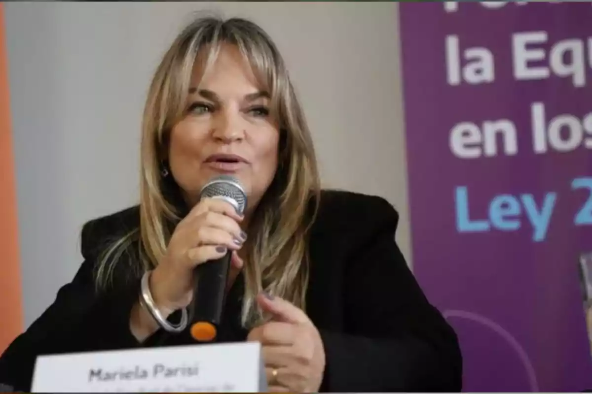 Mujer de cabello rubio hablando por micrófono en un evento con un cartel violeta de fondo Mujer de cabello rubio hablando por micrófono en un evento con un cartel violeta de fondo