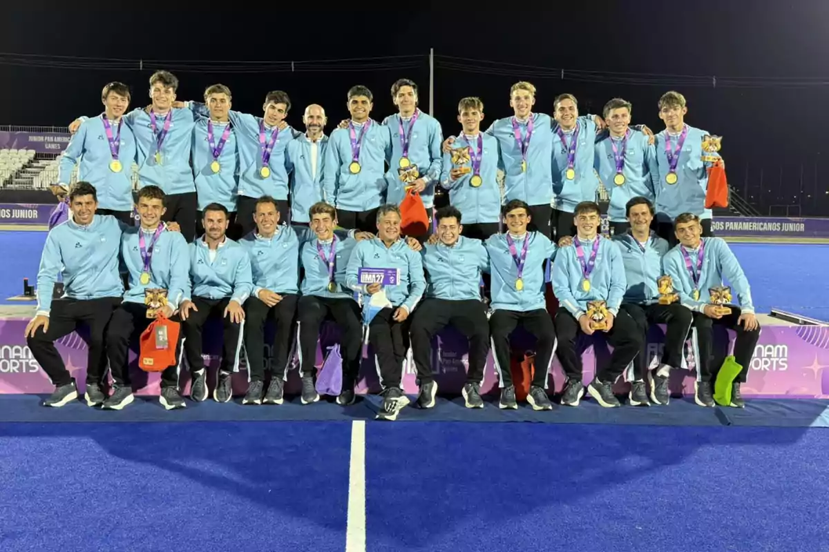 Equipo de jóvenes deportistas con medallas y trofeos posando juntos en una cancha de hockey sobre césped azul, todos vestidos con camperas celestes y pantalones oscuros, celebrando un logro deportivo Equipo de jóvenes deportistas con medallas y trofeos posando juntos en una cancha de hockey sobre césped azul, todos vestidos con camperas celestes y pantalones oscuros, celebrando un logro deportivo