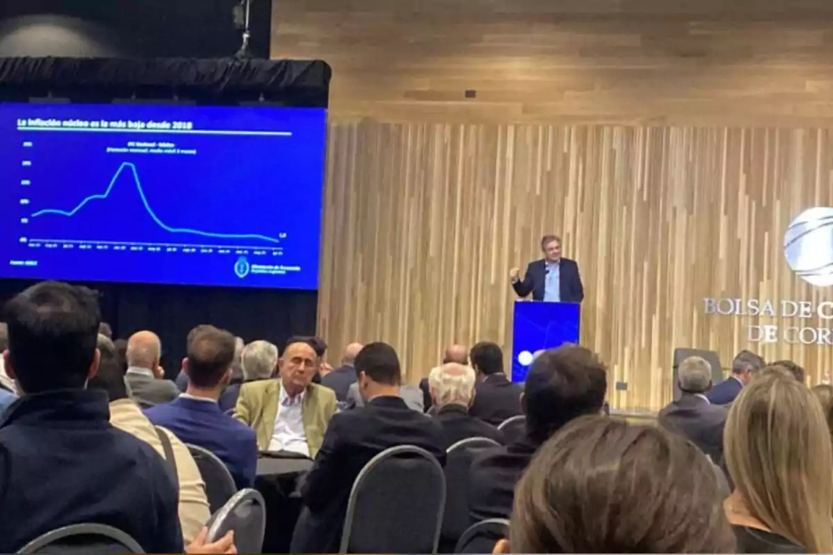 A man gives a lecture in an auditorium full of people while a screen displays a graph about inflation on a blue background.