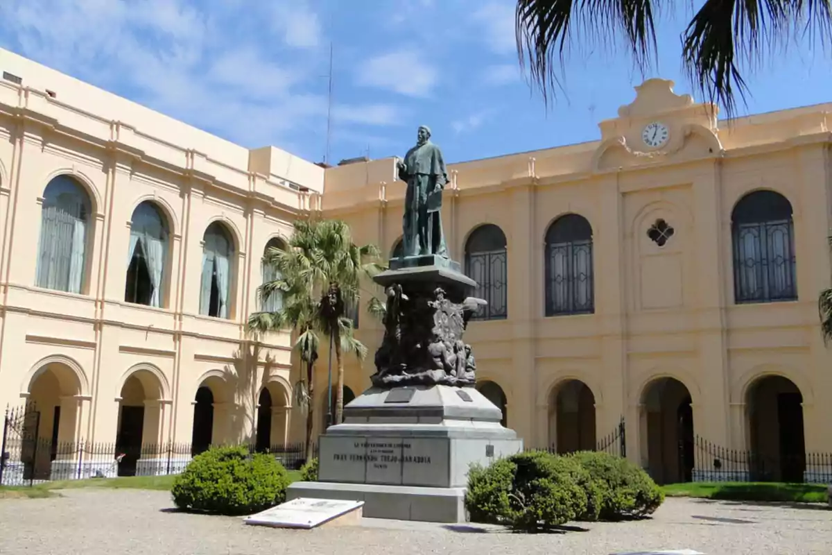 Statue of a historical figure in the center of a courtyard surrounded by a light-colored old building with arched windows and a clock at the top. Statue of a historical figure in the center of a courtyard surrounded by a light-colored old building with arched windows and a clock at the top.