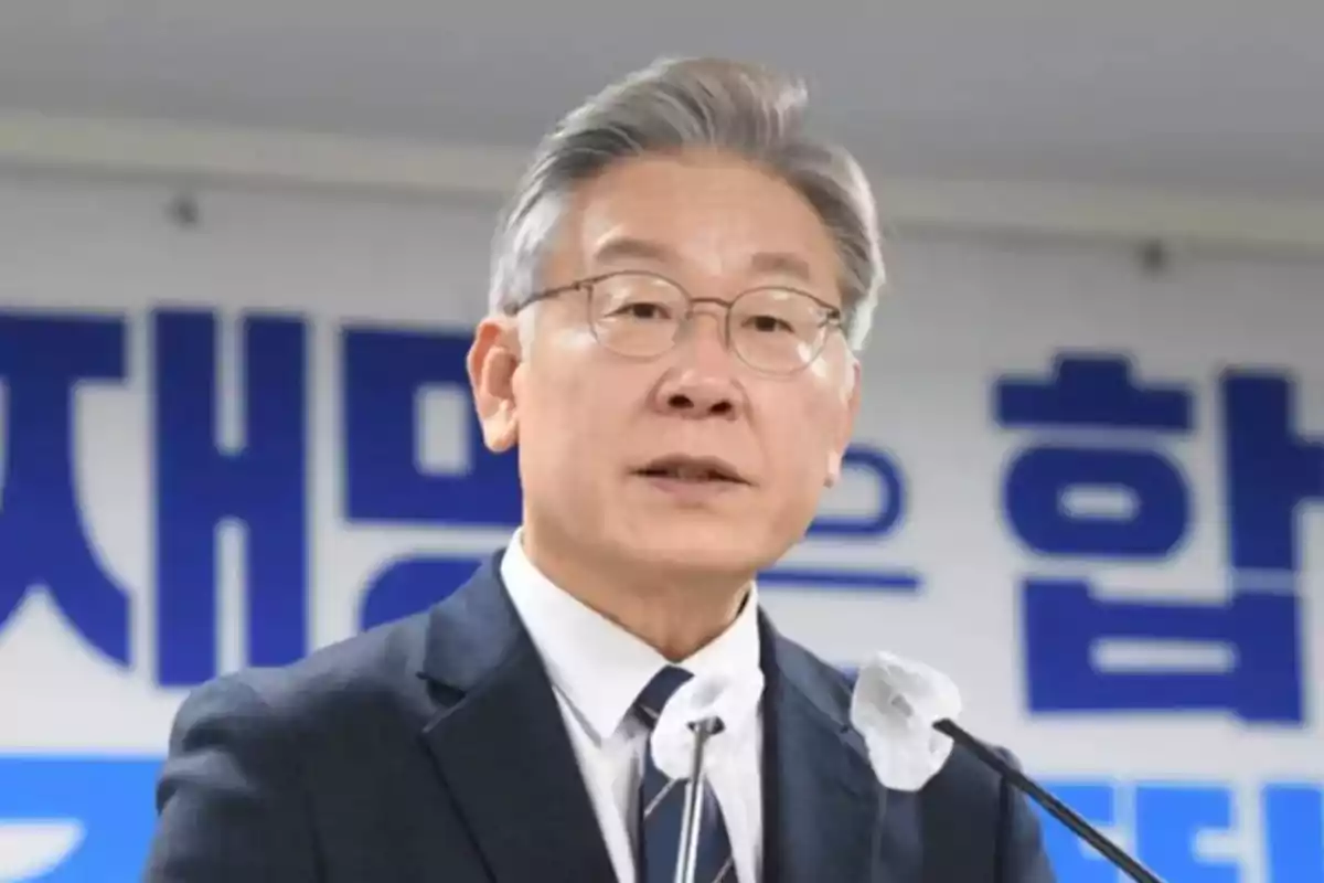Gray-haired man with glasses speaking in front of a microphone with a background of blue Korean letters Gray-haired man with glasses speaking in front of a microphone with a background of blue Korean letters