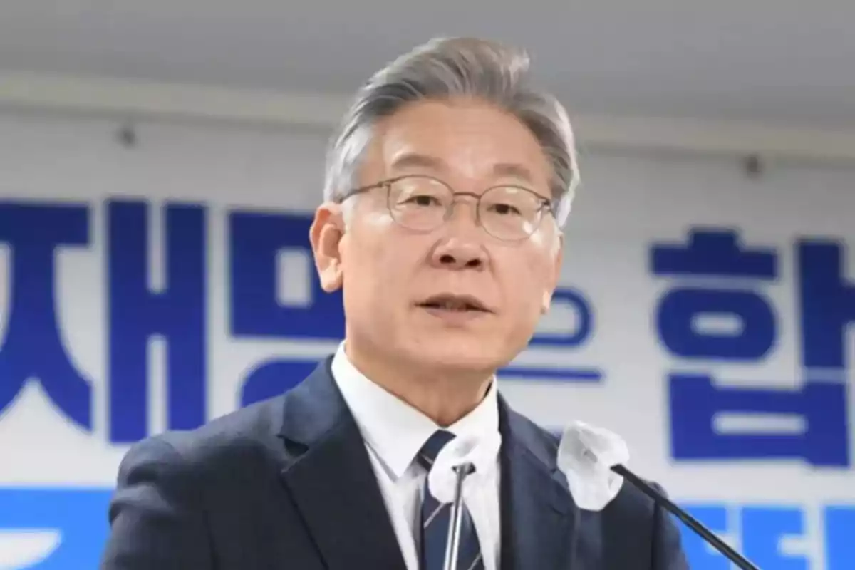 Gray-haired man with glasses speaking in front of a microphone with a background of blue Korean letters Gray-haired man with glasses speaking in front of a microphone with a background of blue Korean letters