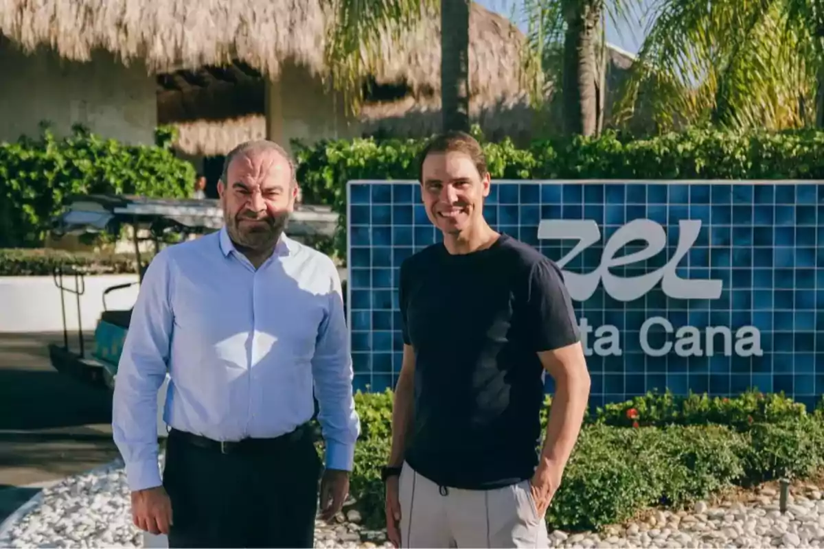 Dos hombres posan sonrientes frente a un letrero azul que dice Zel Punta Cana rodeados de vegetación y palmeras
