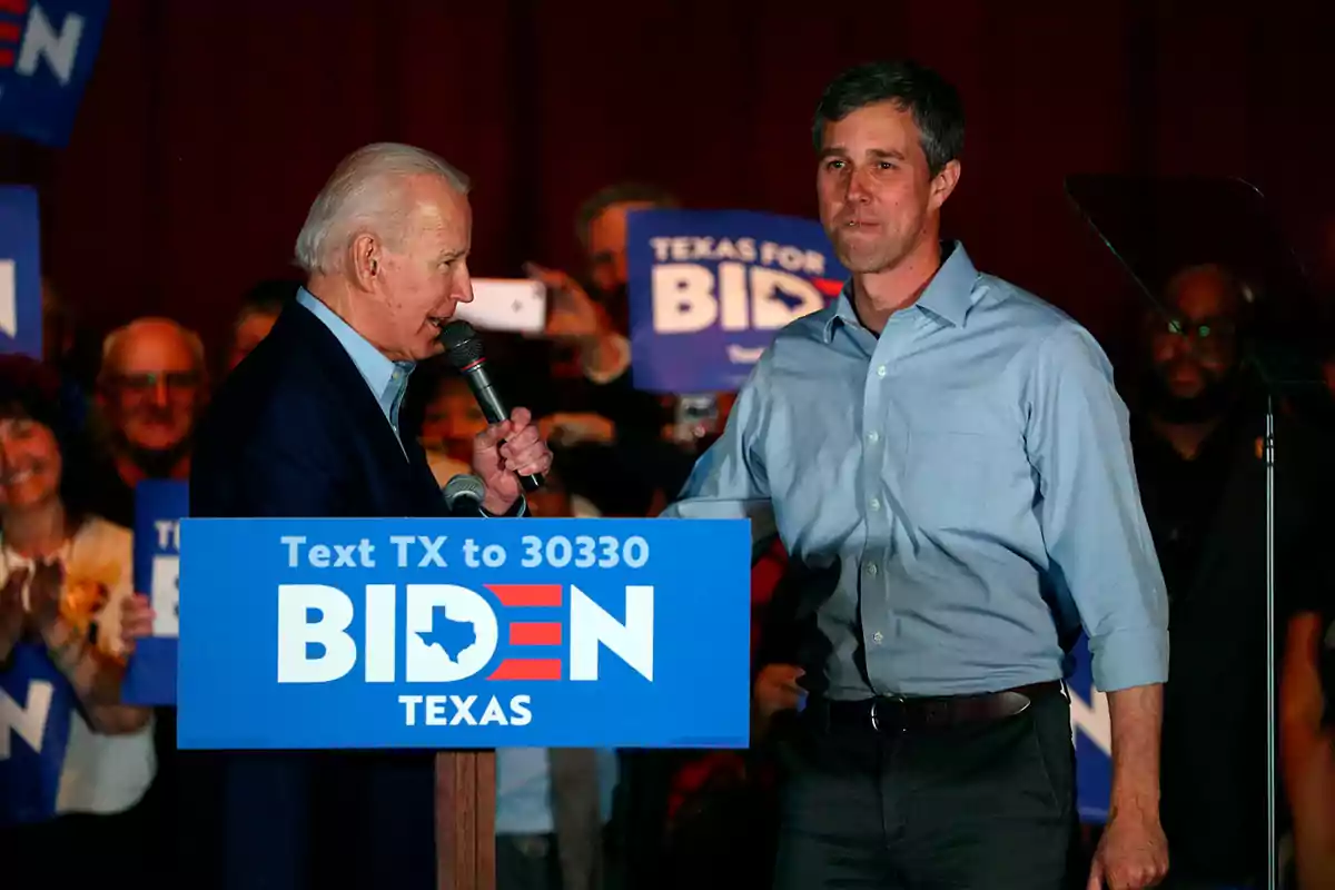 Dos hombres en un mitin político en Texas con un cartel de campaña de Biden y personas aplaudiendo al fondo