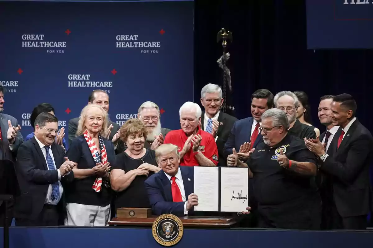 A group of people applauds while a man in the center holds a signed document at an official event with a blue background that says Great Healthcare for You.