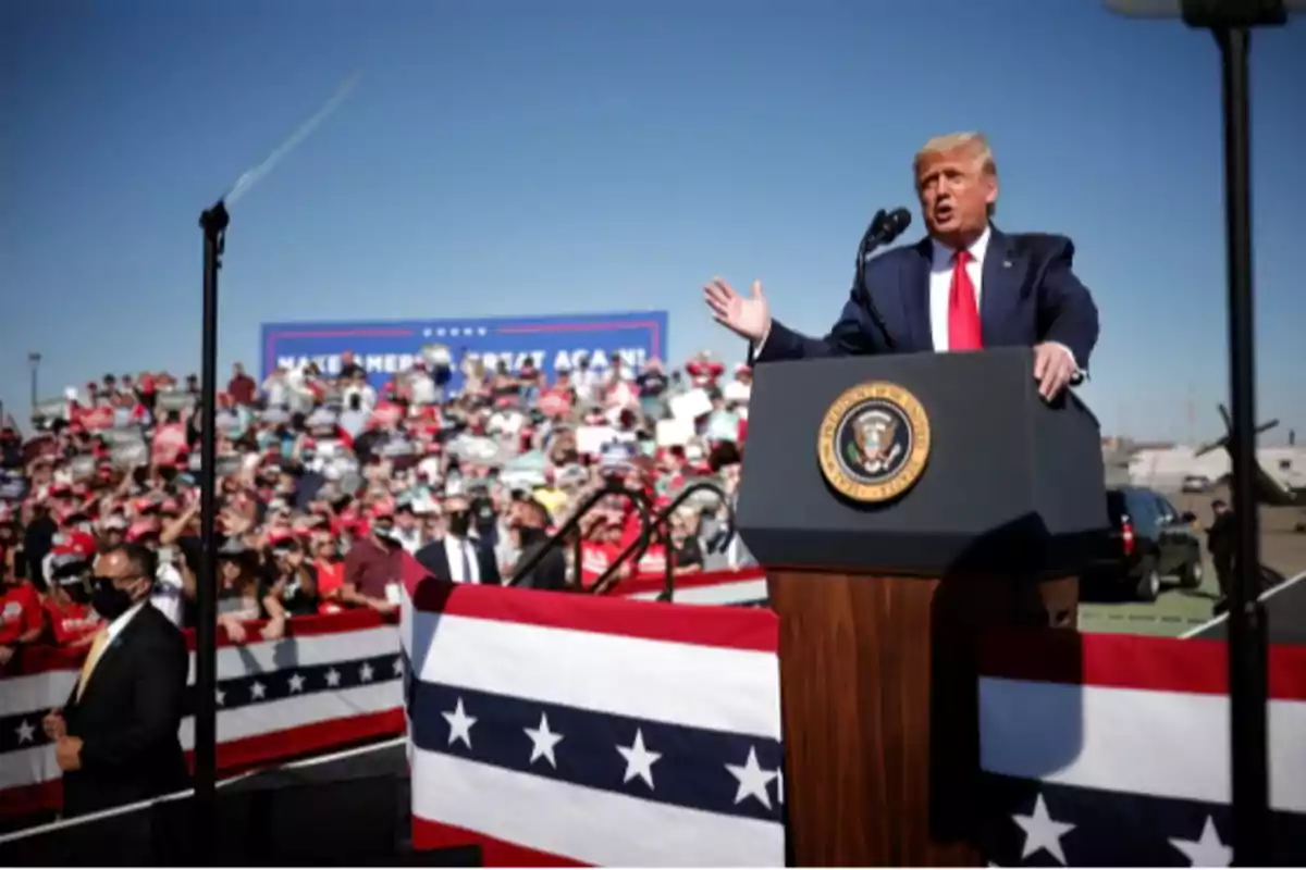 A man in a suit and red tie gives a speech at an outdoor political rally in front of a crowd with flags and patriotic banners.