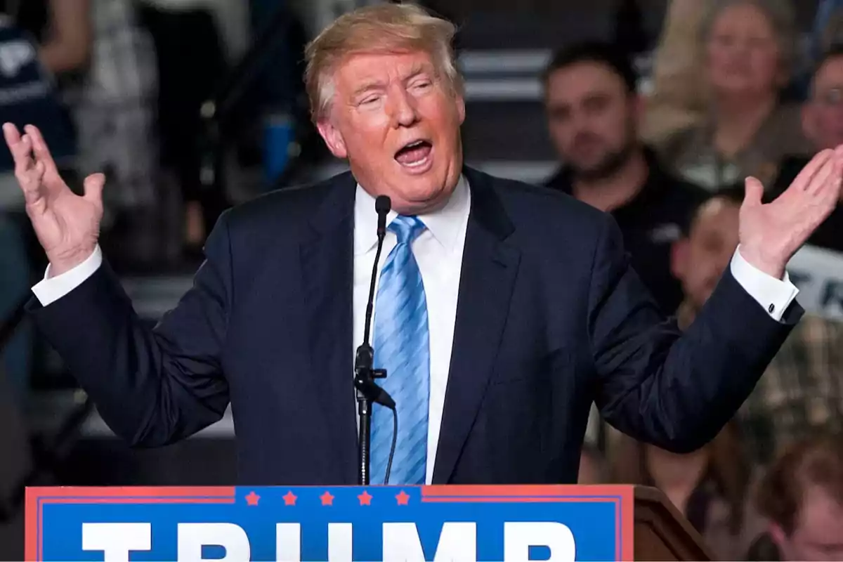 Blond-haired man in a blue suit and light blue tie speaking at a rally in front of a microphone with a sign that says TRUMP and blurred people in the background