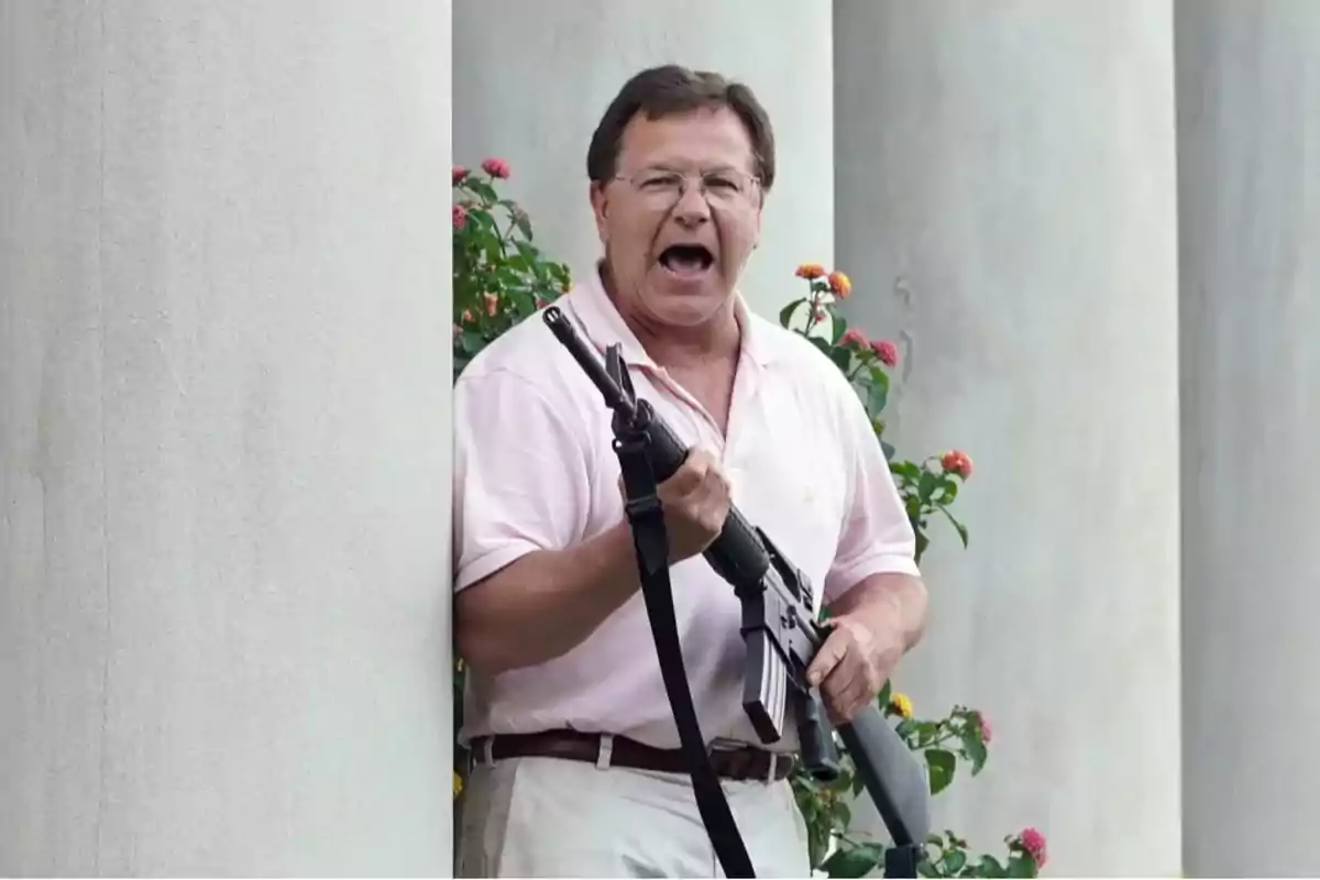 Hombre de mediana edad con camisa polo rosa sostiene un rifle y parece estar gritando mientras está de pie junto a una columna y flores Hombre de mediana edad con camisa polo rosa sostiene un rifle y parece estar gritando mientras está de pie junto a una columna y flores