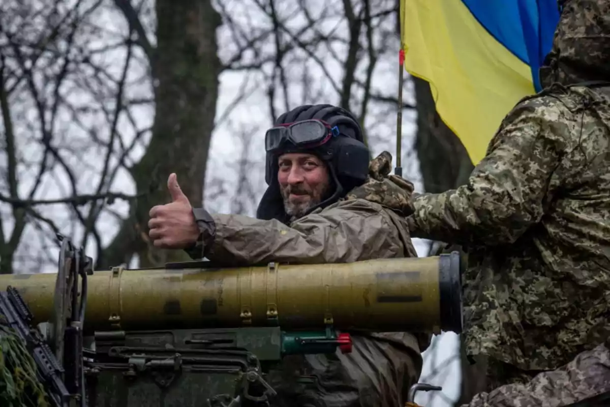 Soldado sonriente con casco y gafas levanta el pulgar mientras está sentado en un vehículo militar junto a una bandera de Ucrania en un entorno boscoso Soldado sonriente con casco y gafas levanta el pulgar mientras está sentado en un vehículo militar junto a una bandera de Ucrania en un entorno boscoso