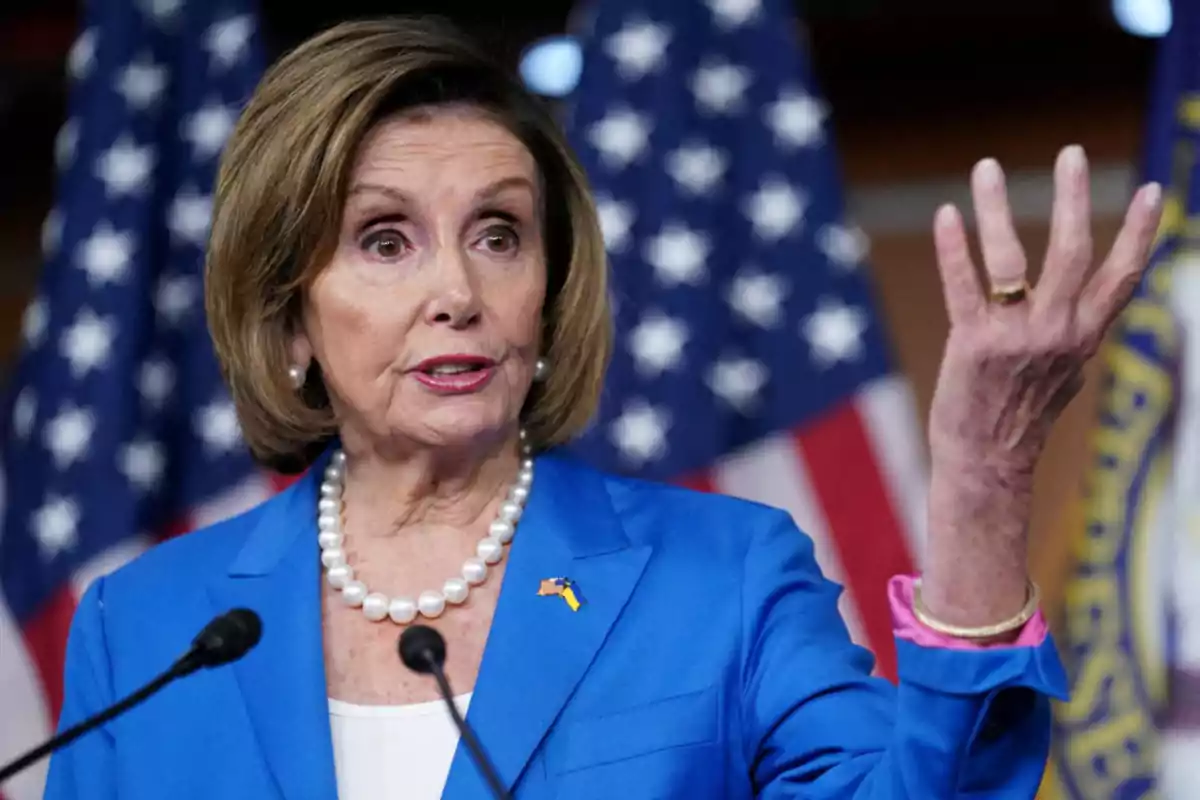 Short-haired blonde woman in a blue blazer speaking in front of microphones with United States flags in the background Short-haired blonde woman in a blue blazer speaking in front of microphones with United States flags in the background