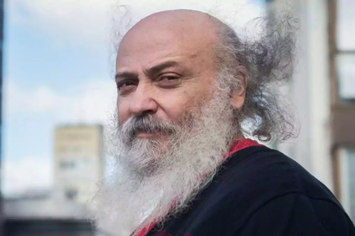Older man with gray hair and beard looking at the camera with a serious expression in a blurred urban setting