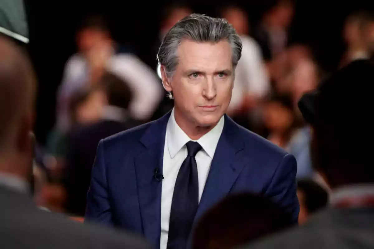 Hombre de cabello canoso con traje azul y corbata oscura en un evento con personas desenfocadas al fondo