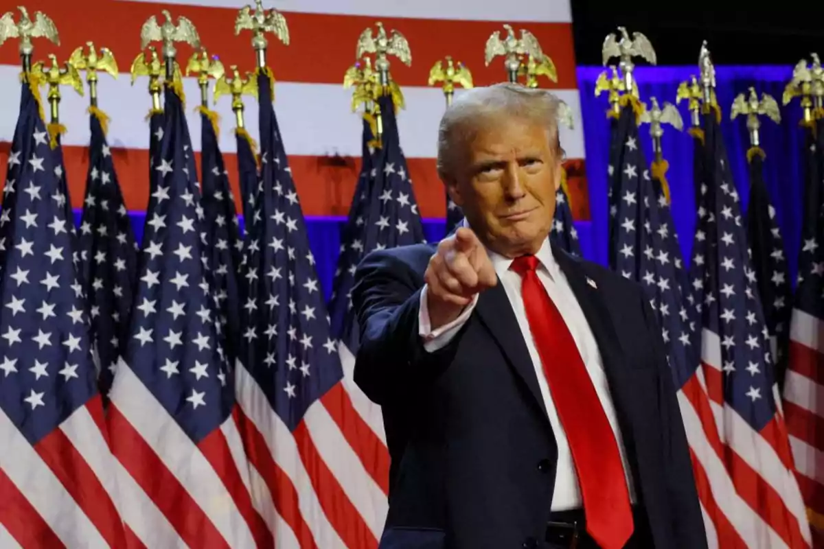 Man in a suit pointing forward with several United States flags in the background