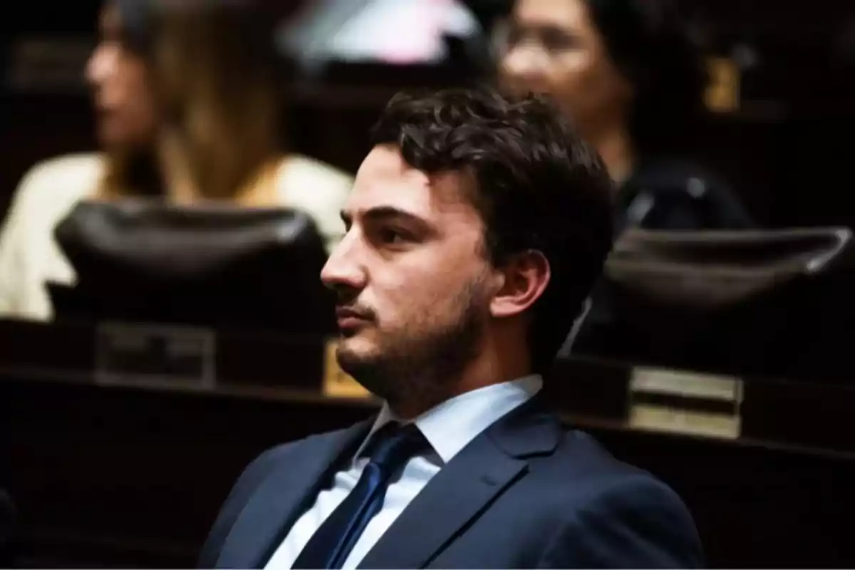 Hombre joven con traje sentado en lo que parece ser un recinto legislativo con otras personas desenfocadas al fondo
