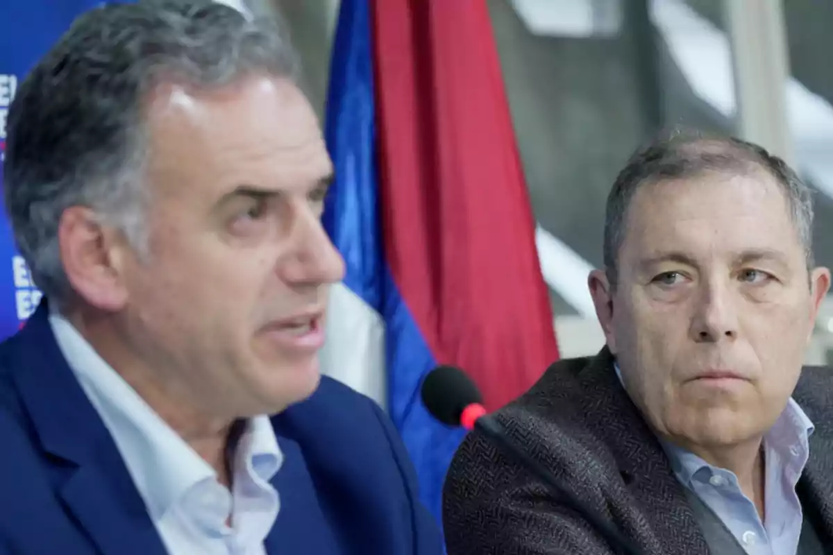 Two men in suits are sitting in front of microphones; one of them is speaking while the other watches him closely, with a flag in the background.