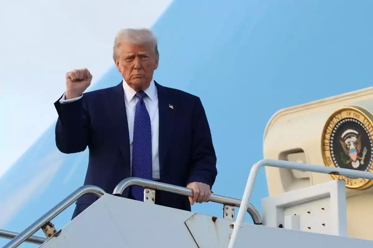 Hombre de traje azul subiendo las escaleras de un avión oficial con el puño levantado y el sello presidencial al fondo