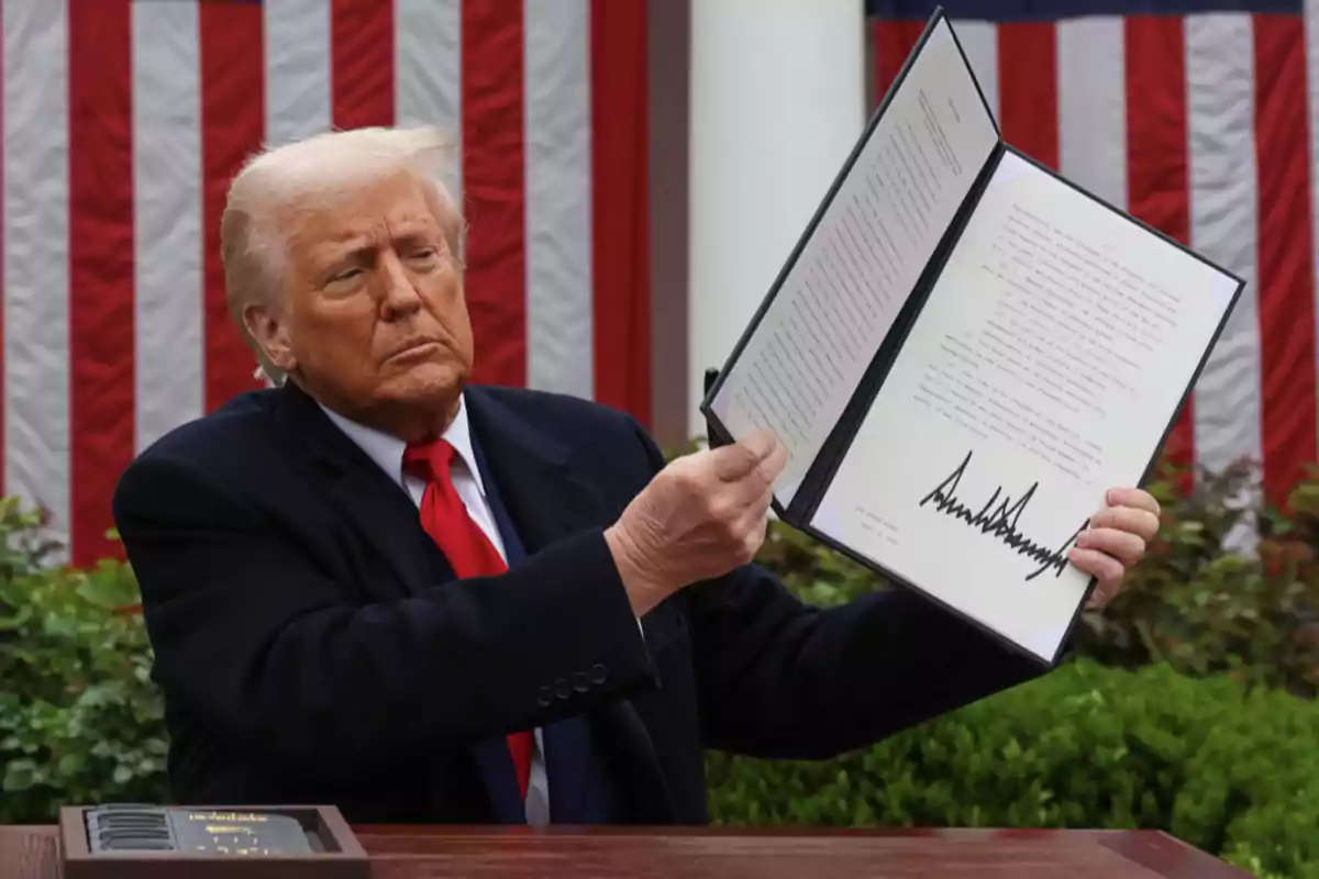 Hombre de cabello rubio con traje oscuro y corbata roja sostiene un documento abierto con una firma grande al aire libre frente a una bandera de Estados Unidos Hombre de cabello rubio con traje oscuro y corbata roja sostiene un documento abierto con una firma grande al aire libre frente a una bandera de Estados Unidos