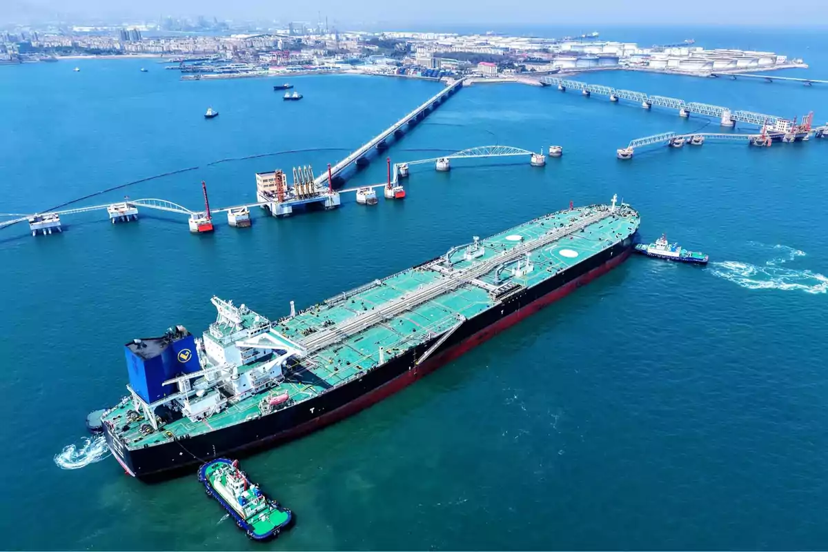 Buque petrolero de gran tamaño siendo asistido por remolcadores en un puerto industrial con infraestructura marítima y tanques de almacenamiento al fondo Buque petrolero de gran tamaño siendo asistido por remolcadores en un puerto industrial con infraestructura marítima y tanques de almacenamiento al fondo