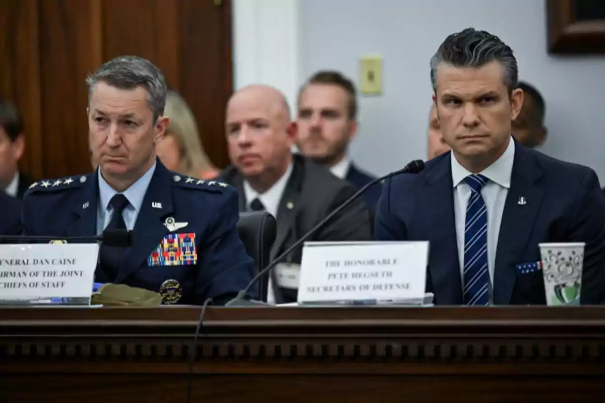 Dos hombres con expresión seria, uno con uniforme militar y el otro con traje, sentados en una mesa durante una audiencia oficial, con varias personas al fondo.