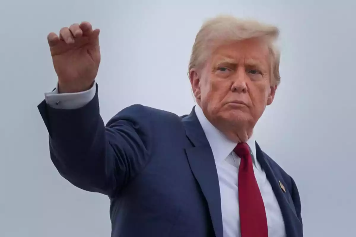 Hombre de cabello rubio con traje azul y corbata roja levantando la mano frente a un fondo gris