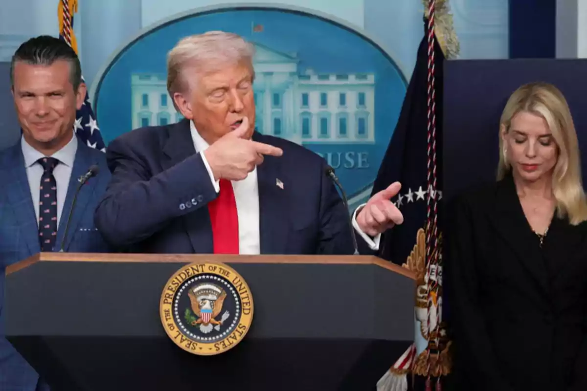 A blond-haired man in a dark suit and red tie gestures with his hand while speaking at a presidential podium, accompanied by a man and a woman.