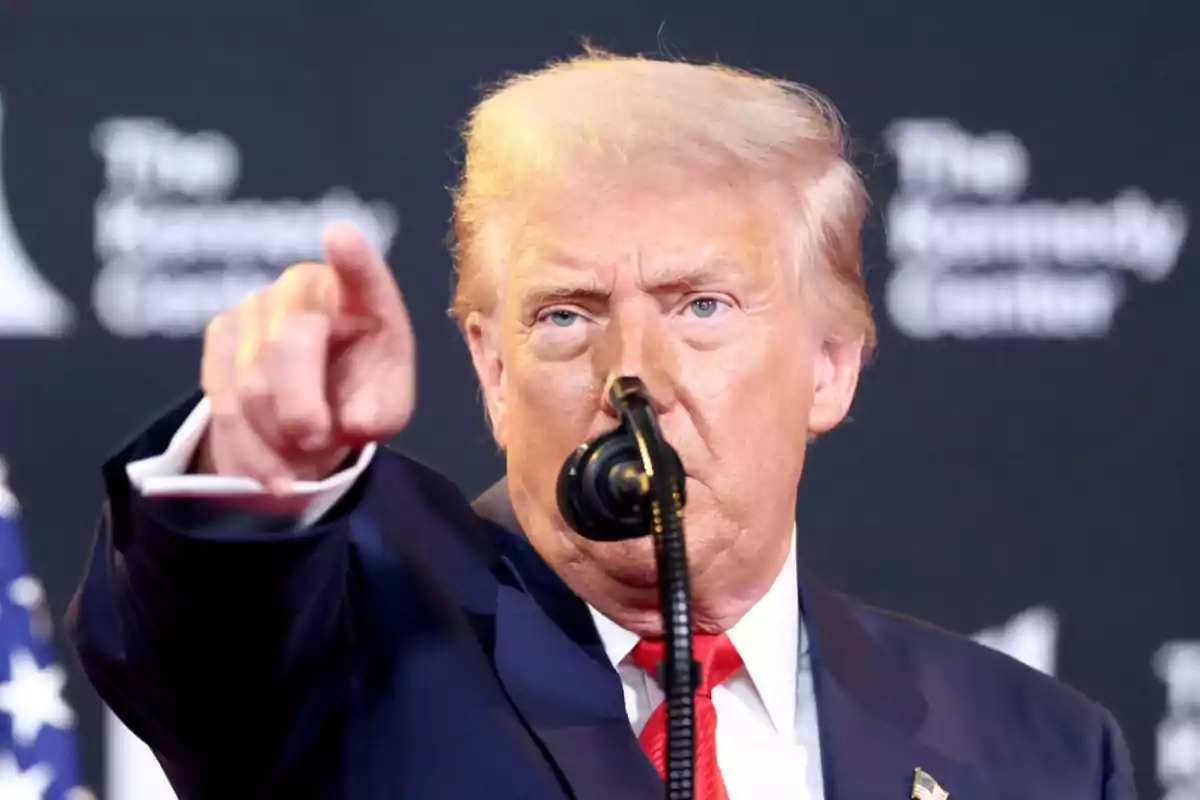 Hombre de cabello rubio con traje oscuro señalando hacia el frente mientras habla en un micrófono Hombre de cabello rubio con traje oscuro señalando hacia el frente mientras habla en un micrófono