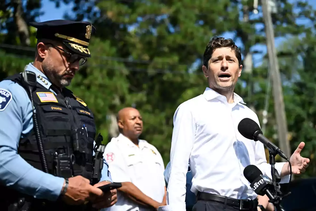 A man in a white jersey speaks in front of several microphones while a police officer and another uniformed person stand beside him in an outdoor setting with trees in the background.