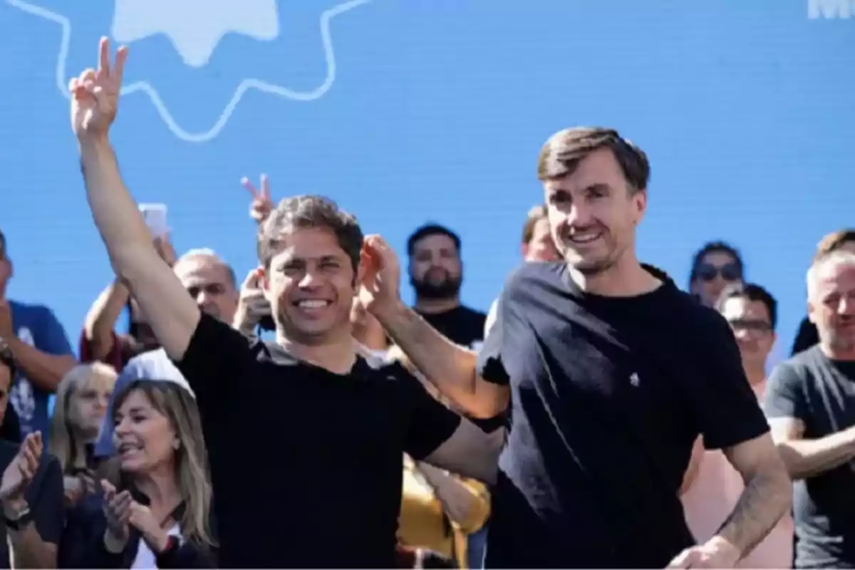Dos hombres sonríen y celebran en un escenario al aire libre rodeados de personas aplaudiendo con un fondo azul