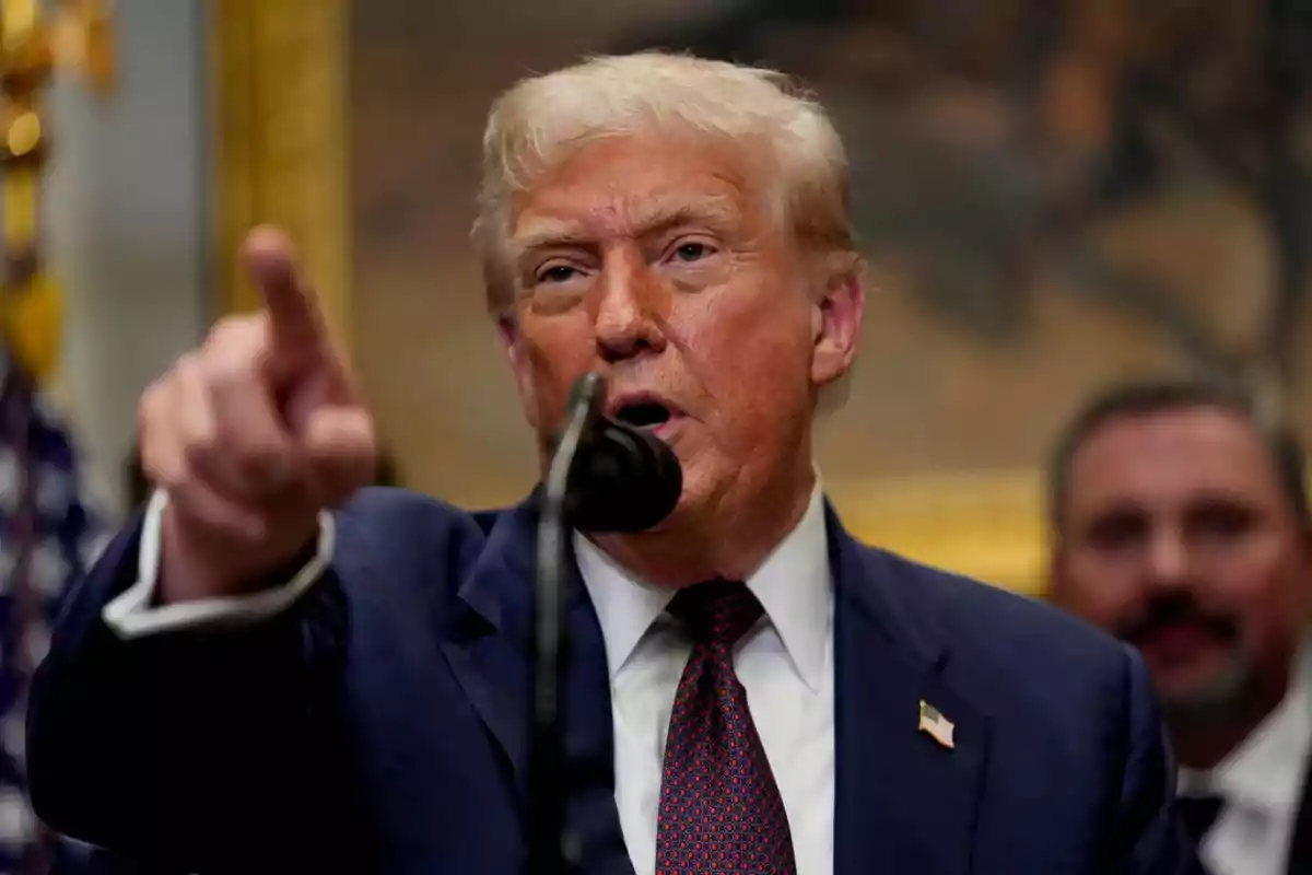 Hombre de cabello rubio con traje azul hablando frente a un micrófono y señalando con el dedo índice Hombre de cabello rubio con traje azul hablando frente a un micrófono y señalando con el dedo índice