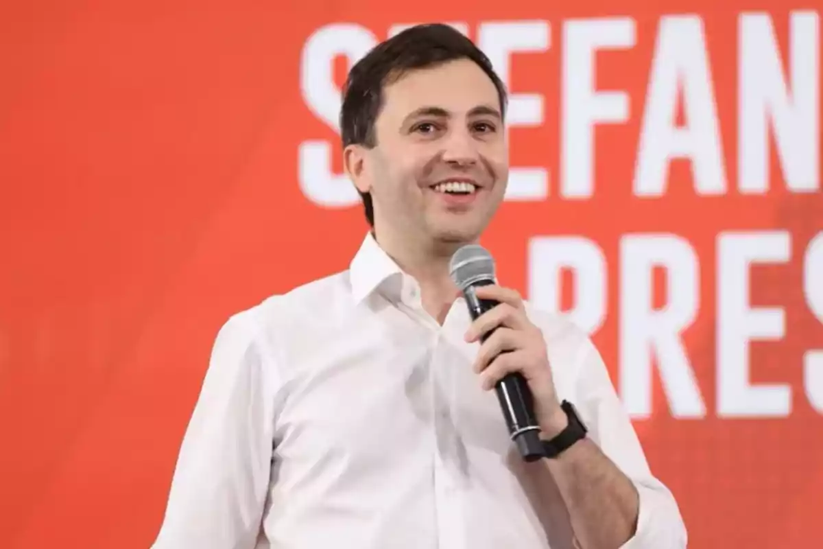 Young man in a white jersey speaking and smiling while holding a microphone in front of a red background with white letters.